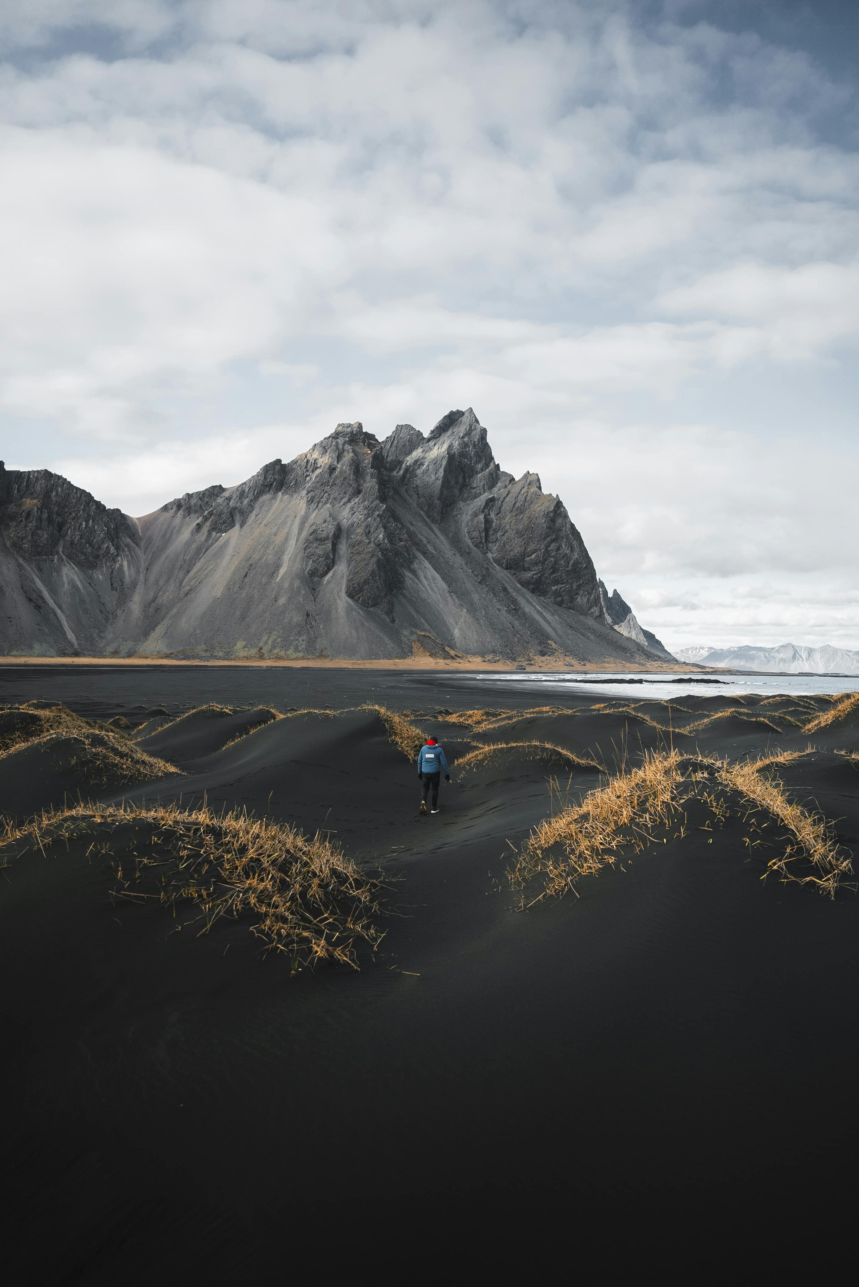 A lone hiker walks through scenic black sand dunes with a stunning Icelandic mountain backdrop under cloudy skies.