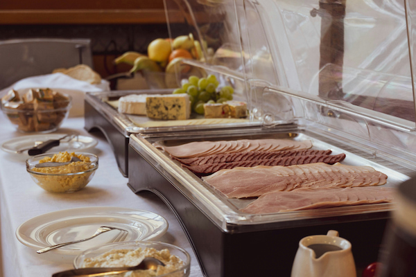 A hotel breakfast buffet display featuring various cold cuts, cheeses, and condiments arranged under clear protective covers on a white tablecloth.