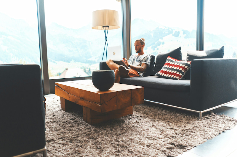A modern lounge area with floor-to-ceiling windows overlooking mountain views. A person sits on a dark sofa using a device, with a wooden coffee table and designer lamp nearby.
